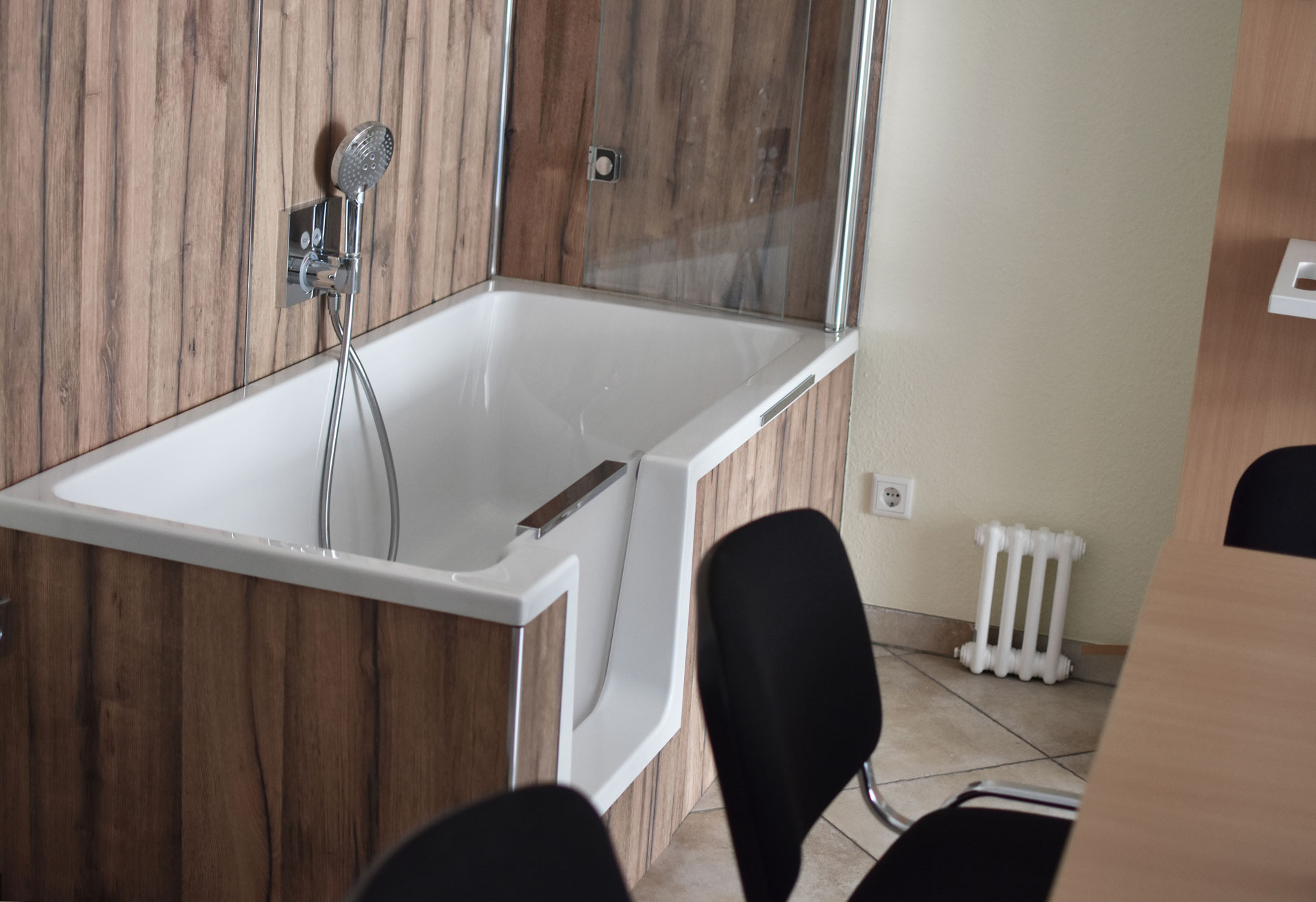 Sitzbadewanne mit Duschfunktion und Holzverkleidung in einem Beratungsraum mit Stühlen und Fliesenboden.