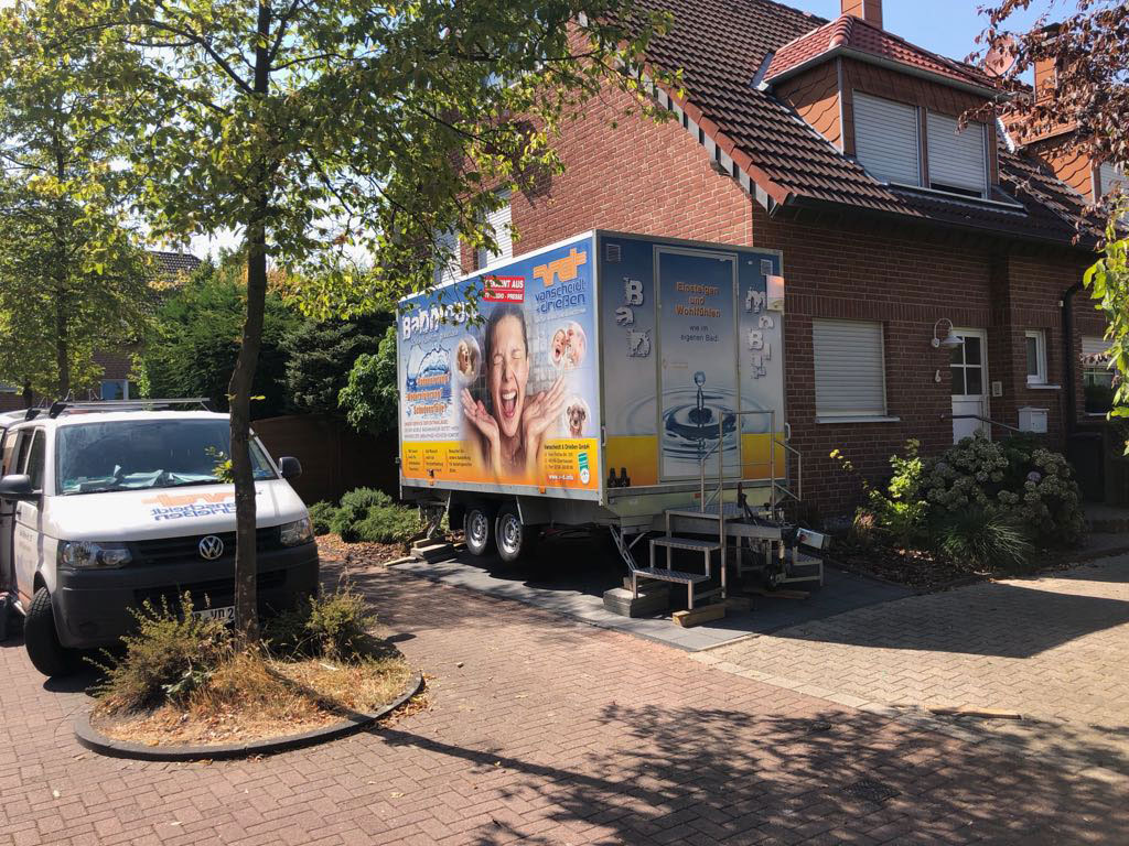 Vor einem Wohnhaus steht das bunte ‚Badmobil‘ der Firma Vanscheidt & Driessen, daneben ein Firmenfahrzeug unter Bäumen.