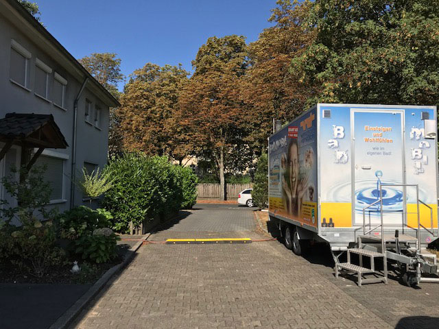 Das mobile Bad ‚Badmobil‘ der Firma Vanscheidt & Driessen steht auf einem Hofplatz mit Rampe, neben Wohnhaus und Bäumen.
